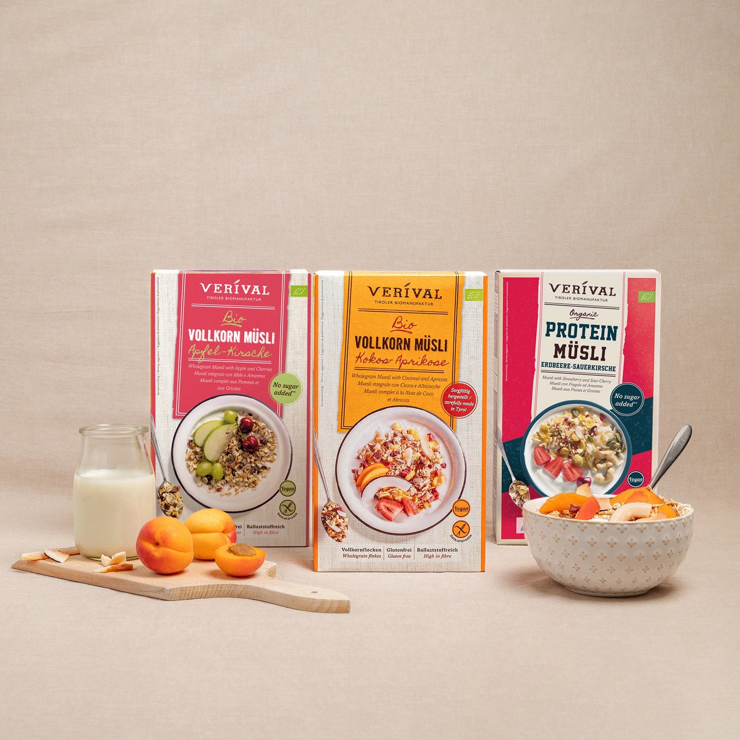 Three boxes of Verival organic wholegrain muesli in various flavors stand upright on a beige surface, next to a bowl of muesli, apple slices, grapes, and a glass of milk.