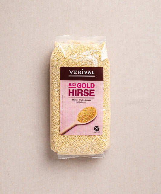 Vor schlichtem Hintergrund steht eine Packung Goldhirse-Hirse der Verival Bio GmbH mit einem Holzlöffel auf dem Etikett. Dieses glutenfreie Produkt ist ballaststoffreich und bietet Gesundheit und Geschmack in jeder Portion.