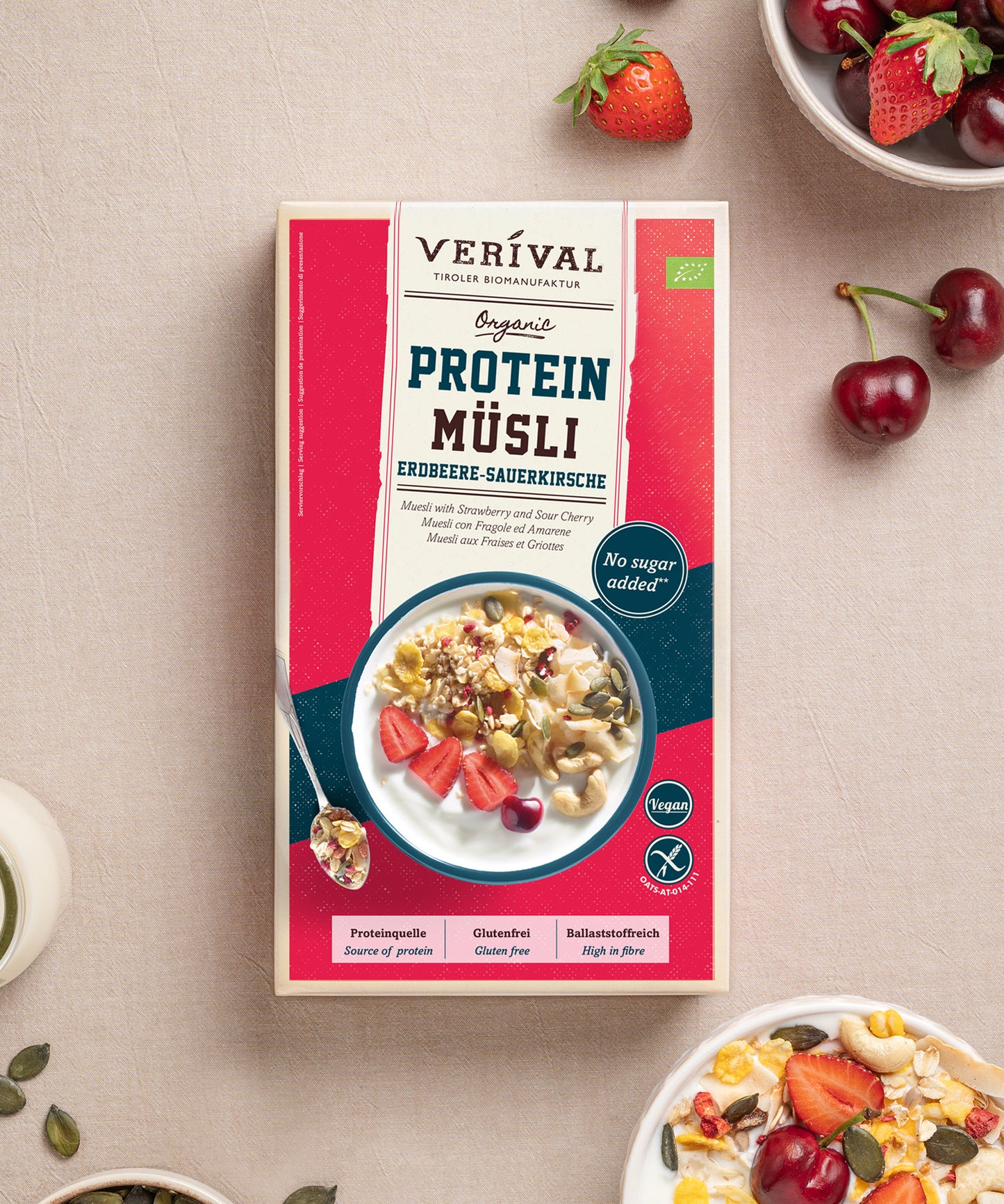 Eine Packung Protein Müsli Erdbeere-Sauerkirsche von Verival Bio wird zusammen mit Müslischalen und frischen Kirschen präsentiert. Es enthält pflanzliche Proteinquelle, ohne Zuckerzusatz, vegan – alles ideal für Sport-Müsli-Enthusiasten.