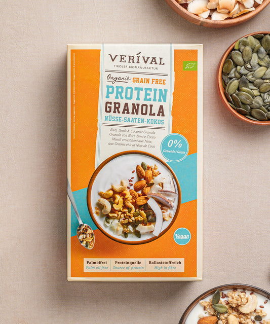 Eine Packung Grain Free Protein-Granola Nüsse-Saaten-Kokos von Verival Bio, reich an pflanzlichen Proteinen, steht auf dem Tisch, umgeben von Schalen mit verschiedenen Nüssen und Samen. Ideal für alle, die ein Low-Carb- oder Sport-Granola suchen, um Energie für den Tag zu tanken.