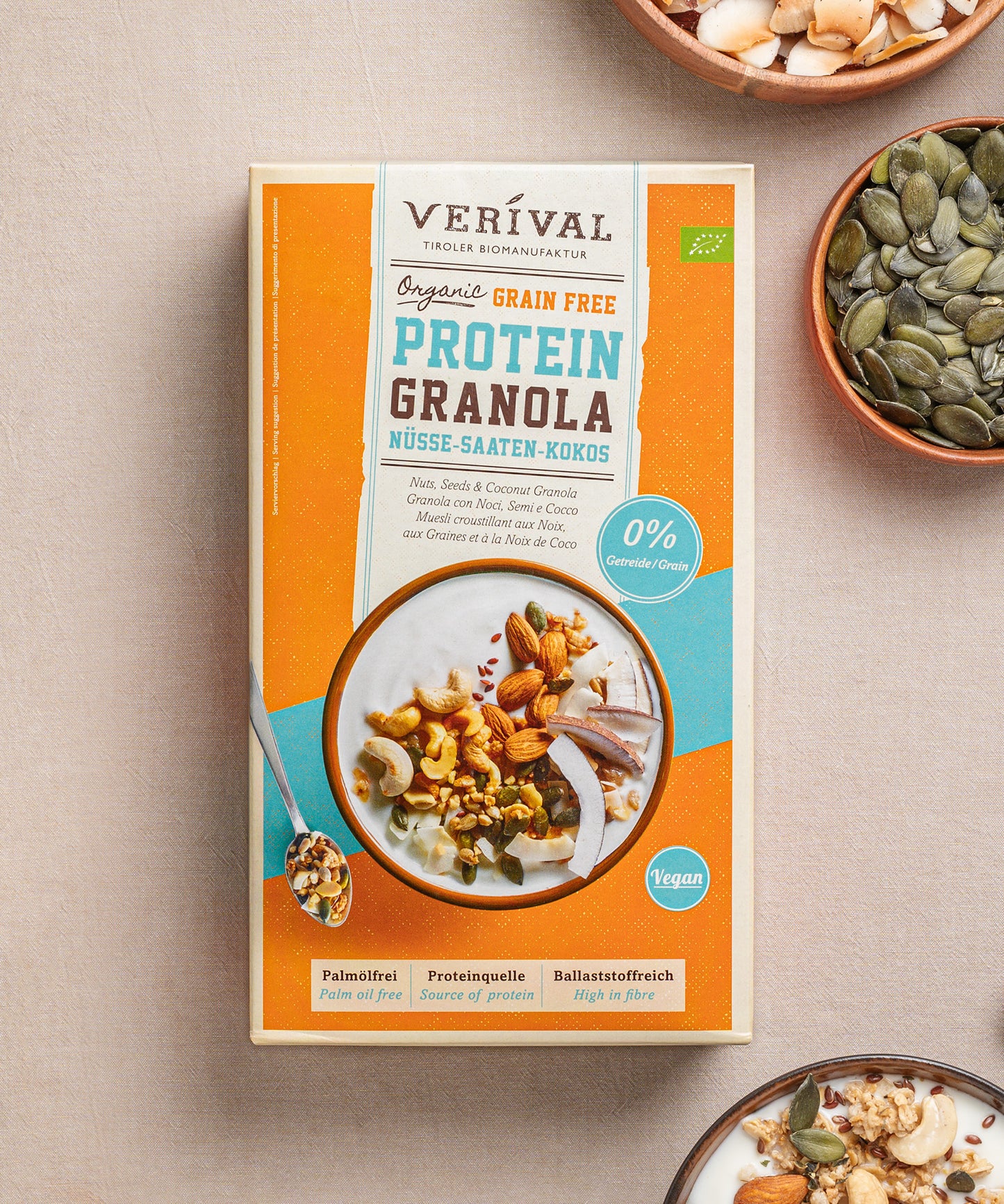 Eine Packung Grain Free Protein-Granola Nüsse-Saaten-Kokos von Verival Bio, reich an pflanzlichen Proteinen, steht auf dem Tisch, umgeben von Schalen mit verschiedenen Nüssen und Samen. Ideal für alle, die ein Low-Carb- oder Sport-Granola suchen, um Energie für den Tag zu tanken.