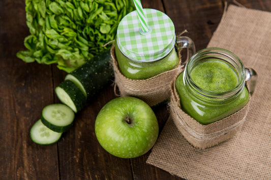 Zwei Einmachgläser mit grünem Smoothie auf Sackleinen, mit grünem Apfel, Gurkenscheiben und Blattsalat auf einem Holztisch.
