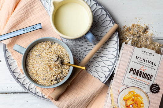 Eine Schüssel Porridge mit Löffel, ein Krug Milch, eine Packung Verival Aprikosen-Honig-Porridge und etwas verschütteter Porridge auf einem Tablett mit Serviette.