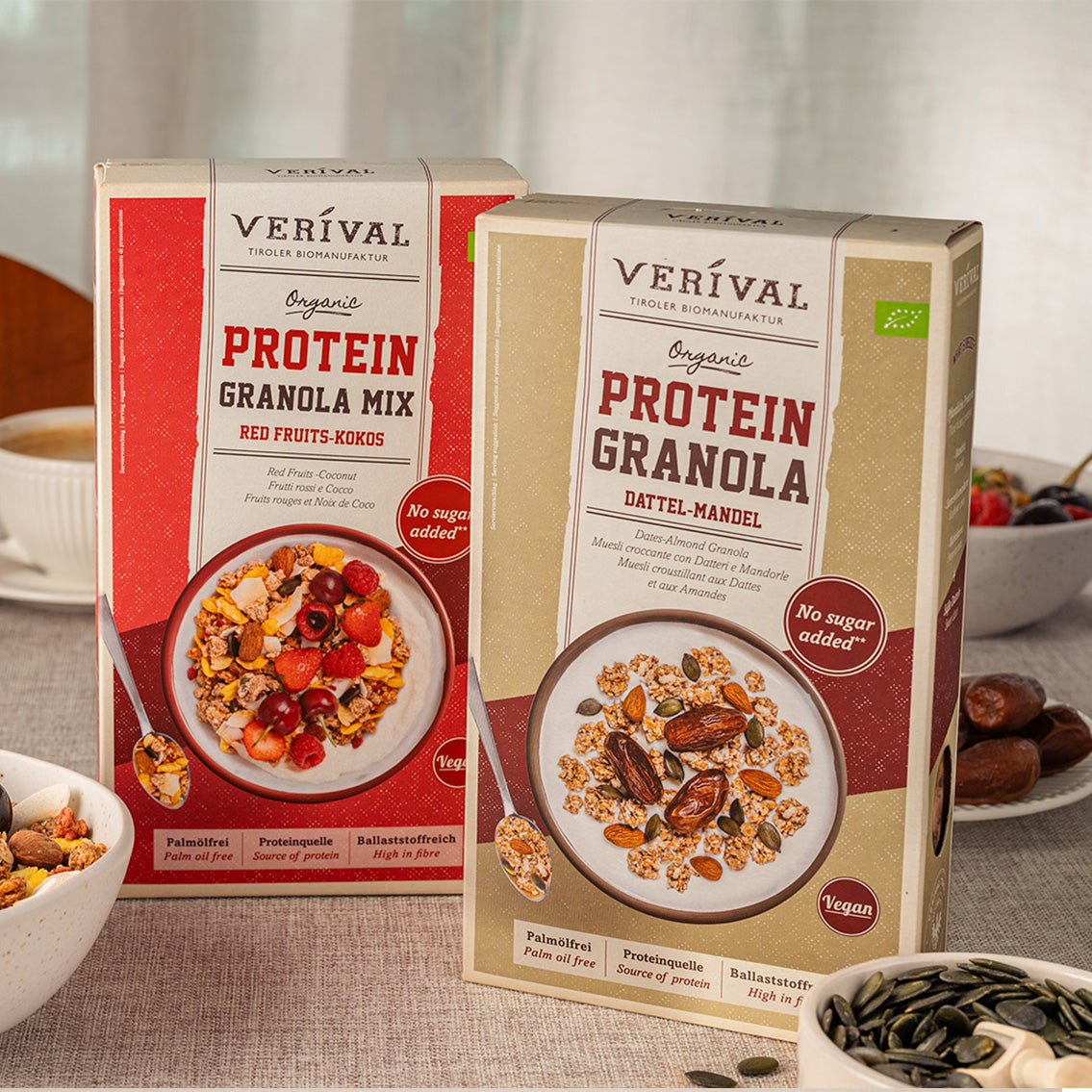 Zwei Schachteln Verival Bio-Protein-Granola, eine mit roten Früchten und Kokosnuss und eine mit Datteln und Mandeln, stehen auf einem Tisch mit Granola-Schalen und Zutaten.