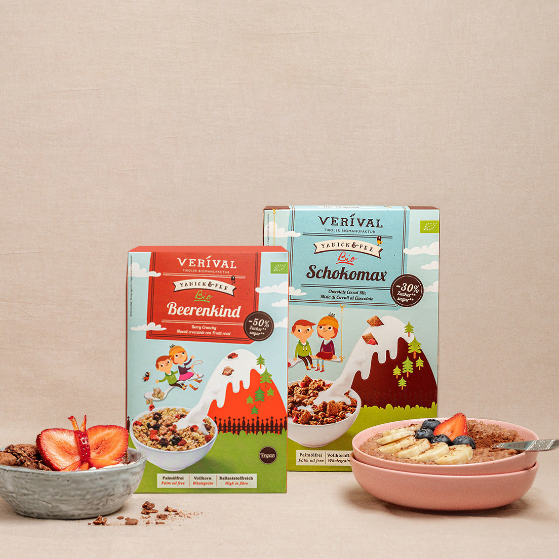 Two packs of Verival muesli, Beerenkind and Schokomax, stand upright next to a muesli bowl with strawberry and banana slices on a beige background.