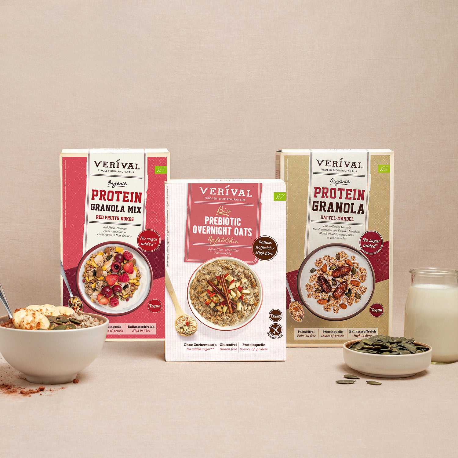 Three packages of Verival breakfast cereal, including Protein Granola Mix, Prebiotic Overnight Oats, and Protein Granola. In front are a bowl of muesli, a spoon, nuts, and a glass of milk.