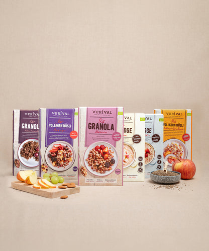 Fünf Schachteln des Bestsellers „Probierpaket“ der Verival Bio GmbH stehen nebeneinander und präsentieren verschiedene Müslisorten. Im Vordergrund steht auf einer beigen Unterlage ein Schneidebrett mit Apfelscheiben, Weintrauben und einer Schale mit Chiasamen.