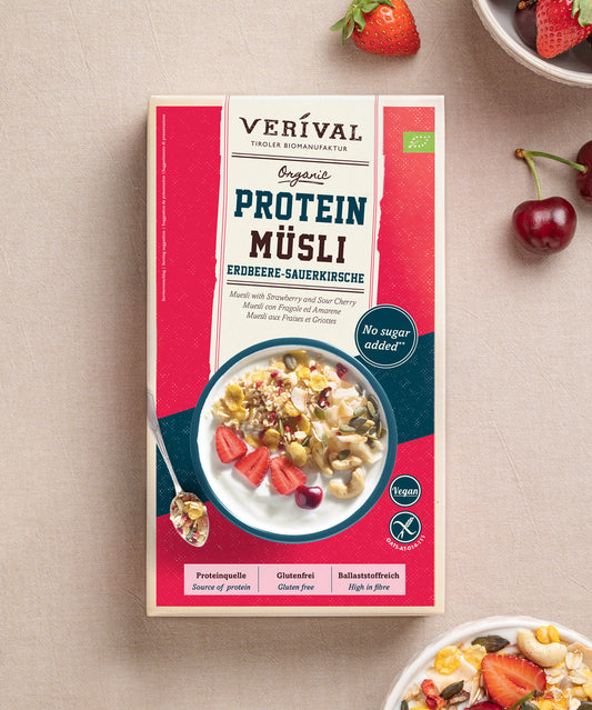 Eine Packung Protein Müsli Erdbeere-Sauerkirsche der Verival Bio GmbH steht auf einem hellen Untergrund mit frischen Kirschen und einer Schale Sport Müsli. Diese pflanzliche Proteinquelle wird ohne Zuckerzusatz hergestellt und sorgt für einen natürlich leckeren Start in den Tag.