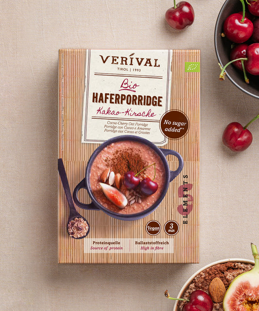 Eine Dose Verival Bio GmbH Bio Haferporridge Kirsche-Kakao steht auf einer beigen Fläche mit Kirschen, Mandeln und einem Löffel. Die Verpackung betont ballaststoffreich, vegan und ohne Zuckerzusatz - ideal für ein warmes Frühstück.