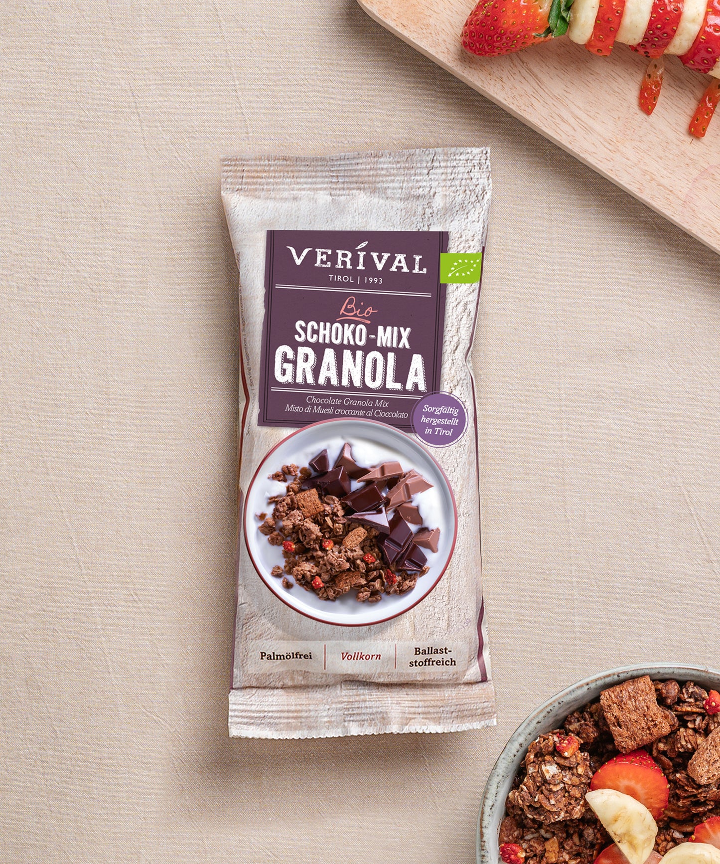 Eine Packung Bio Schoko Mix Granola der Verival Bio GmbH steht auf einer beigen Fläche neben einer Schüssel mit knusprigen Frühstücksflocken und einem Holzbrett mit Obstspießen - perfekt für ein ballaststoffreiches Frühstück.