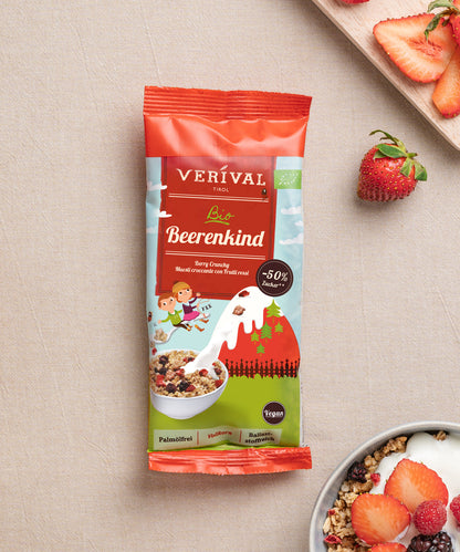 Eine Packung Verival Bio GmbH Bio Beerenkind Beerenmüsli liegt auf einer beigen Fläche neben einer Schale veganem Crunchy Frühstück mit Milch und geschnittenen Erdbeeren.