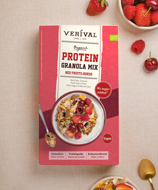 Verpackung von Verival Bio Bio Red Fruits Protein Granola Mix mit roten Früchten und Kokosnuss, darauf eine Schüssel mit dem ballaststoffreichen Müsli, vermischt mit Joghurt und einem Löffel, umgeben von frischen Beeren und Kirschen.