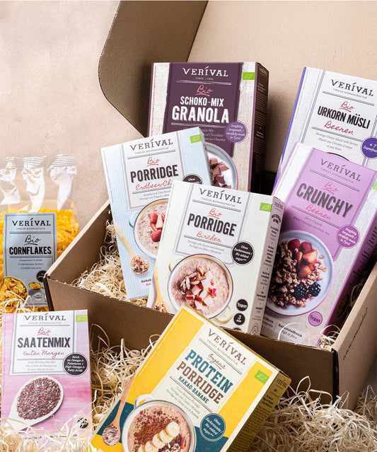 Das Set "Best Breakfast Friends" der Verival Bio GmbH zeigt eine Auswahl an Frühstücksflocken - Granola, Porridge, Müsli, Cornflakes und Körnermischung - ansprechend auf Stroh neben einer offenen Pappschachtel arrangiert.