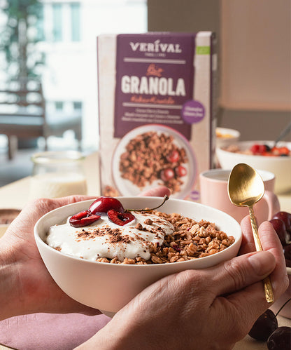 Eine Person hält eine Schüssel „Verival Bio Kakao-Kirsche Granola glutenfrei“ mit Joghurt und Kirschen in der Hand und hat einen goldenen Löffel in der Hand. Im Hintergrund hebt eine Verpackung der Verival Bio GmbH in einer sanft beleuchteten Essensszene die veganen und glutenfreien Eigenschaften des Produkts hervor.