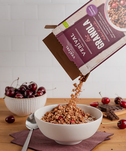 Eine Hand schüttet „Verival Bio GmbH Bio Kakao-Kirsche Granola glutenfrei“ in eine weiße Schüssel. Daneben liegen ein silberner Löffel, eine Serviette, Kirschen und Schokoladenstückchen – vor dem Hintergrund einer weißen Backsteinwand ergibt sich so ein verlockendes glutenfreies Frühstück.
